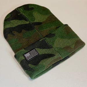 May include: A green camouflage beanie with a folded brim. The beanie features a small embroidered American flag patch on the front. The hat is made of a knit fabric.