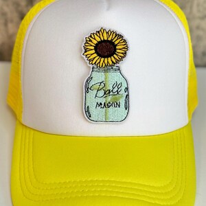 May include: A bright yellow and white trucker hat featuring an embroidered patch on the front. The patch depicts a yellow sunflower inside a light blue glass Ball Mason jar with the text Ball Mason clearly visible on the front of the jar.