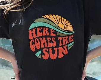 May include: Black t-shirt with a retro-style graphic of a sun rising over a wave. The graphic says "Here Comes The Sun" in a colorful, vintage font.