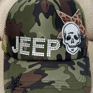 May include: Camouflage baseball cap with the word "JEEP" spelled out in rhinestones. The cap features a skull patch, a leopard print star, and a black beaded star. The mesh back is olive green.