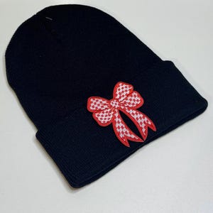 May include: Black beanie with a red and white checkered bow applique. The beanie is made of a soft knit material and is a stylish accessory for cold weather. The bow adds a decorative touch.