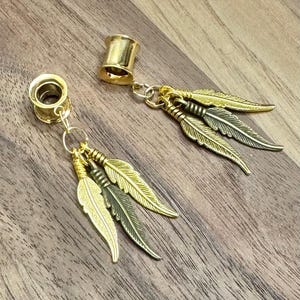 May include: Gold-colored ear gauges with dangling feather charms. The gauges are cylindrical with a flared edge. Each charm features three detailed feather pendants in gold and bronze tones, connected by small rings.