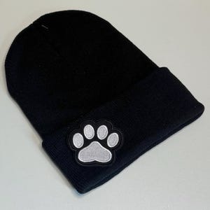 May include: Black knit beanie with a folded brim. A white paw print patch with a black border is sewn on the front. The hat is made of a soft, warm material, perfect for cold weather.