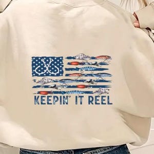 May include: A beige sweatshirt with a graphic of an American flag made of fishing lures and the text "Keepin' It Reel".