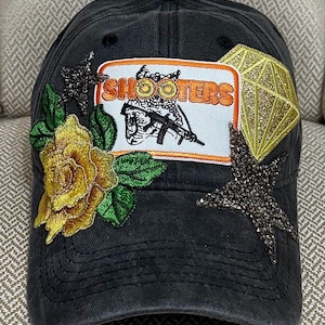 May include: Black baseball cap with embroidered gold rose, green leaves, and a patch that says "SHOOTERS" with a graphic of a person holding a gun. The cap is embellished with a gold diamond and two star-shaped rhinestones.