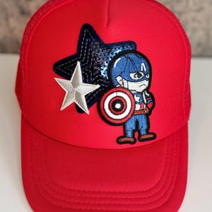May include: Red baseball cap featuring a Captain America design. The cap has a mesh back and a curved brim. The front of the cap displays an embroidered Captain America character, a white star, and a blue sequined star.