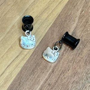 May include: A pair of black ear gauges with dangling silver-colored Hello Kitty charms. The charms feature the iconic cat face with a bow. The gauges and charms are displayed on a wooden surface.