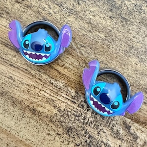 May include: Two blue and purple Stitch-themed phone grips. Each grip features a smiling Stitch face with large black eyes, a dark blue nose, and purple ears. The grips are attached to silver metal rings.
