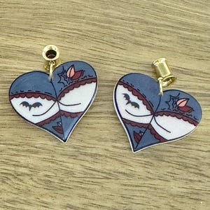 May include: A pair of heart-shaped earrings with a blue, white, and burgundy design. Each earring features a bat, spiderweb, and floral accents. The earrings have gold-colored posts.