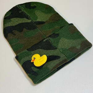 May include: Green camouflage beanie with a yellow rubber ducky patch. The beanie is made of a knit fabric and features a folded cuff. The duck patch has an orange beak and eye.