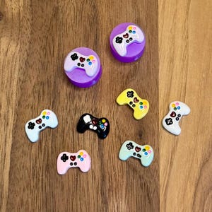 May include: A collection of colorful video game controller charms in various colors, including purple, black, pink, yellow, and blue. The charms are arranged on a wooden surface, showcasing their vibrant details and playful design.