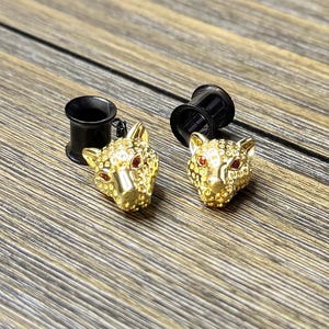 May include: A pair of ear plugs featuring gold-toned leopard head charms with textured surfaces and small red inset stones for eyes. Each leopard head is attached to a smooth, black cylindrical ear tunnel designed for stretched ear piercings.