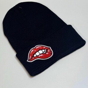 May include: Black knit beanie with a red lips patch. The patch features a red outline and a white set of teeth. The beanie is made of a soft, knitted material and is suitable for cold weather.