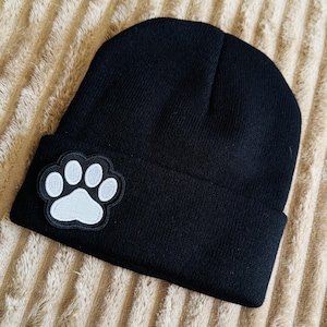 May include: A black knit beanie featuring a folded cuff. Attached to the front of the cuff is a circular embroidered patch displaying a white paw print design with a black border. The hat is constructed from a solid black fabric material.