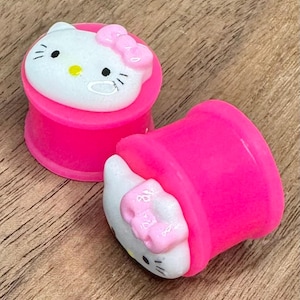 May include: Two bright pink ear gauges with a white and pink Hello Kitty design. The Hello Kitty face has black eyes and whiskers, with a pink bow on top. The gauges are on a wooden surface.