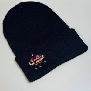 May include: Black knit beanie with a folded cuff. The beanie features an embroidered patch of a pink, yellow, and white UFO with small yellow lights. The hat is made of a soft, knitted material.
