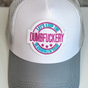May include: White and gray trucker hat with a mesh back and a curved brim. The front panel features a circular patch with the text "LET'S KEEP THE DUMBFUCKERTY FUN TODAY" in pink and blue embroidery.