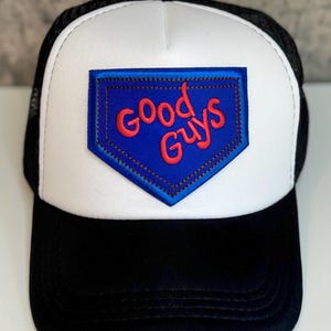 May include: Black and white trucker hat with a white front panel and a black mesh back. The front features a blue patch with the words "Good Guys" in red lettering.