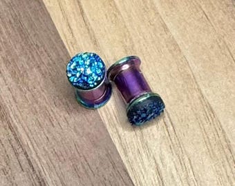May include: Two iridescent purple and blue ear gauges with a sparkling blue top. The gauges are cylindrical with a flared edge. The gauges are made of metal and have a glittery, textured surface.