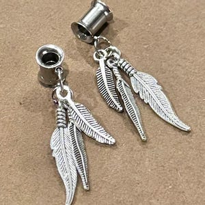 May include: Two silver-toned ear gauges with dangling feather charms. The gauges are cylindrical with a flared edge, and the feathers are detailed with etched lines. The jewelry is displayed on a neutral, light brown surface.