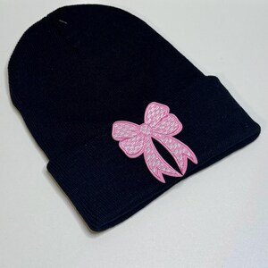 May include: A black beanie hat with a pink bow applique. The bow is detailed with a textured pattern and outlined in pink. The hat is made of a soft, knitted material, perfect for cold weather.
