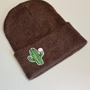 May include: A brown knit beanie with a green cactus patch featuring a pink flower. The beanie is made of a soft, ribbed knit material. The cactus patch is detailed with white outlines.