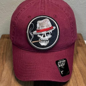 May include: A maroon baseball cap featuring a circular embroidered patch on the front. The patch depicts a skull wearing a fedora with a red band and smoking a cigarette. A black sticker with the text OTTO is attached to the lower right brim.