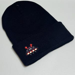 May include: Black beanie with a folded brim. The beanie features a colorful embroidered patch of a UFO with red, white, and purple accents. The hat is made of a soft, knitted material.