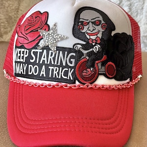 May include: A red and white trucker hat with a red rose, a rhinestone star, and a black and red embroidered image of a character from the movie Saw. The hat has a chain trim and the text "Keep Staring I May Do A Trick" on the front.