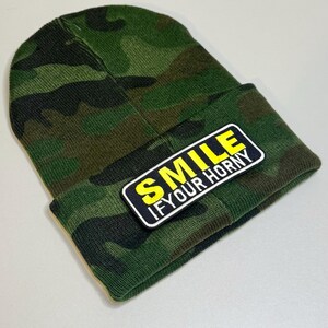 May include: A green camouflage beanie with a folded brim. A rectangular patch with the words "SMILE IF YOUR HORNY" in yellow and white lettering is sewn onto the brim. The beanie is made of a soft, knitted material.