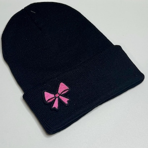 May include: A black knit beanie with a folded brim. A pink bow patch is sewn on the front. The beanie is made of a soft, textured material, perfect for cold weather. This hat is a stylish accessory.