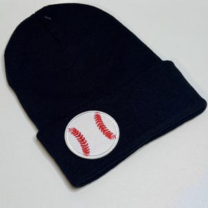 May include: A black beanie hat with a white baseball patch. The baseball patch features red stitching details. The hat is made of a soft, knitted material. This hat is a great accessory for baseball fans.