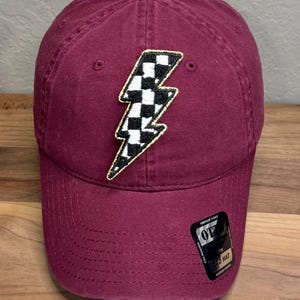 May include: A maroon baseball cap featuring a central embroidered lightning bolt patch with a black and white checkered pattern and a gold border. A black rectangular sticker with the text OTTO DAD HAT is attached to the lower right brim.