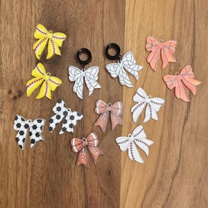 May include: A collection of sports-themed bow earrings and plugs. The bows feature designs of softballs, soccer balls, footballs, and baseballs in various colors. The earrings are displayed on a wooden surface.