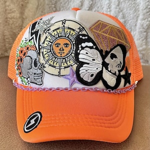 May include: Orange and white trucker hat with various colorful patches, including a compass, a sun, a butterfly, a skull, a diamond, and a lightning bolt.