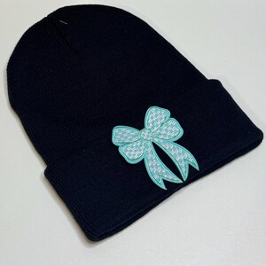May include: Black beanie hat with a light blue bow applique. The bow features a white and light blue houndstooth pattern. The hat is made of a knitted material and is folded at the brim.