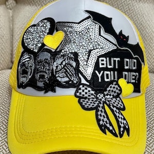 May include: Yellow and white trucker hat with a curved brim. The hat features a star, heart, and bat embellishments, along with the phrase "BUT DID YOU DIE?" and a checkered bow. The hat has a mesh back.