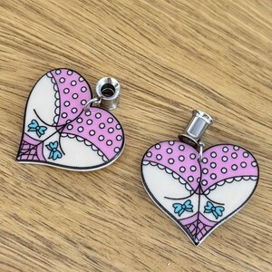 May include: Two heart-shaped ear gauges with a pink polka dot design and white accents. Each gauge features a silver-colored post. The design includes blue bows and a black outline, creating a playful aesthetic.