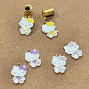 May include: Gold-colored ear gauges with dangling white Hello Kitty charms. The charms feature a white body, a bow, and accents in yellow, pink, and purple. The charms are enamel-coated and have a gold-colored loop.