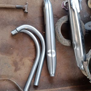 Puede incluir: Varios componentes de escape de motocicleta se muestran en una superficie de trabajo. Las piezas incluyen tubos curvos y dos silenciadores plateados. También se ven un martillo y una llave. La imagen incluye el texto "OPPO A12 • ©Iwan custom 2023/05/21 10:00."