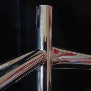 May include: Close-up of a polished chrome bicycle frame with three intersecting tubes. The frame is shiny and reflects light.