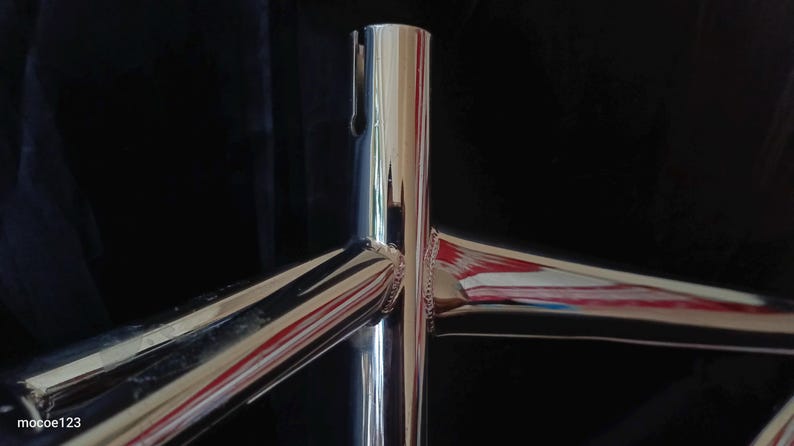 May include: Close-up of a polished chrome bicycle frame with three intersecting tubes. The frame is shiny and reflects light.