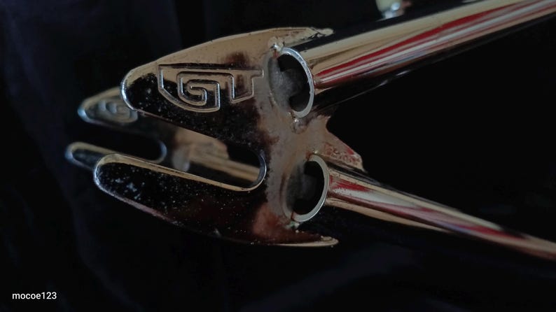 May include: Close-up of a chrome bicycle frame with the word "GT" embossed on the top tube.