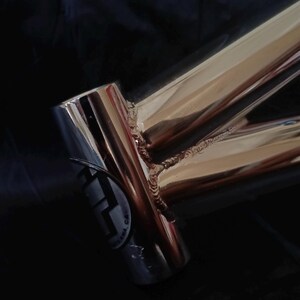 May include: Close-up of a polished chrome bicycle frame with a welded joint. The frame has a sticker with the text "F.A.M.A. CA."