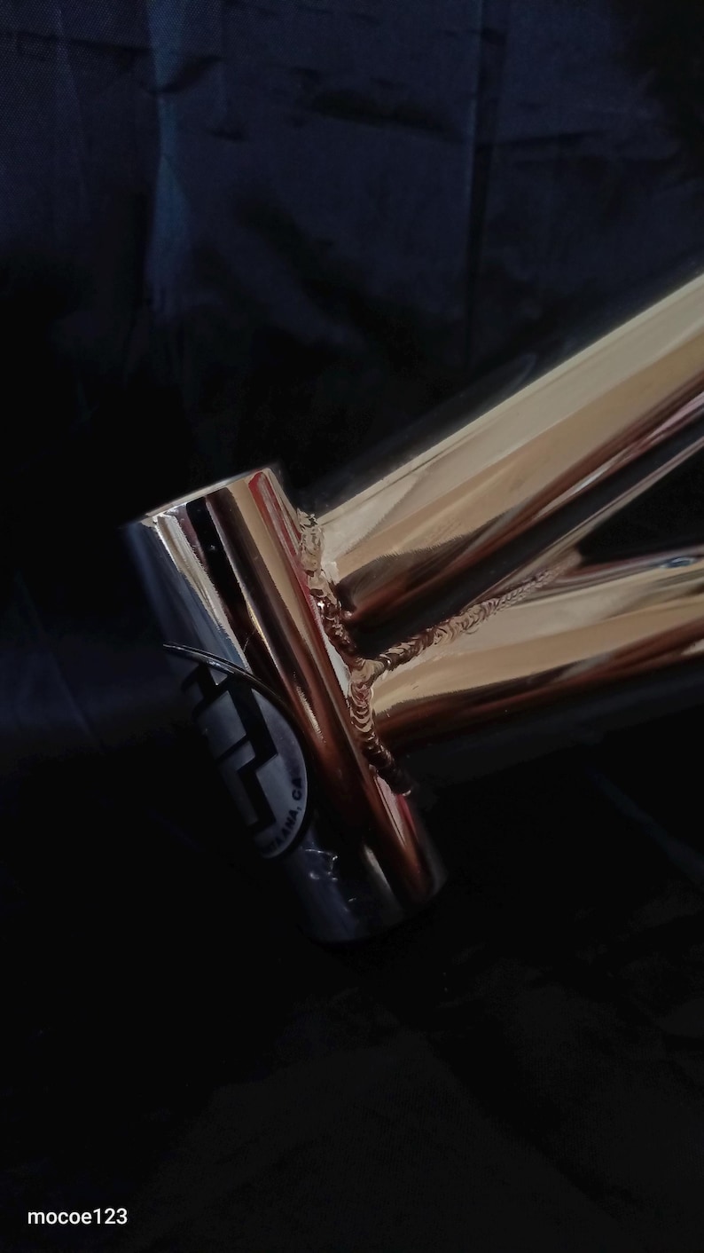 May include: Close-up of a polished chrome bicycle frame with a welded joint. The frame has a sticker with the text "F.A.M.A. CA."