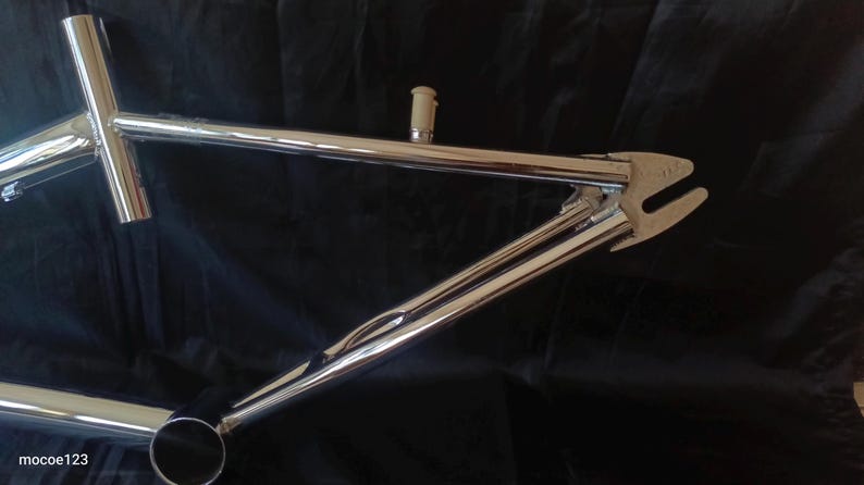 May include: Chrome bicycle frame with a silver finish. The frame has a curved top tube and a straight down tube. The frame is designed for a BMX bike.