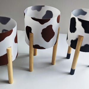 May include: Three white ceramic planters with brown, black and grey cow print designs. Each planter is on a wooden stand with black painted tips.