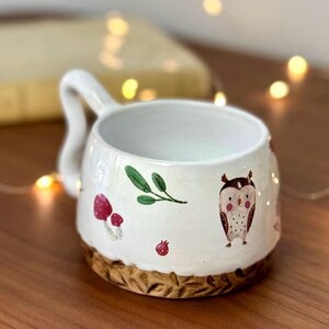 May include: White ceramic mug with a brown rim and a hand-painted design of an owl, mushrooms, and leaves.