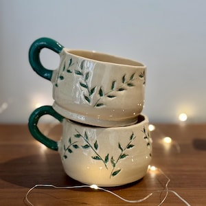 Puede incluir: Dos tazas de cerámica apiladas con asas verdes y un diseño de hojas. Las tazas son de color beige claro con hojas verdes pintadas en los lados.
