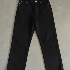 Peut inclure: Un pantalon en jean noir uni doté d'une taille haute, d'une fermeture à bouton et à glissière, de deux poches avant et de deux petites poches gousset. Le jean présente une coupe droite avec des rivets et des boutons en métal argenté.